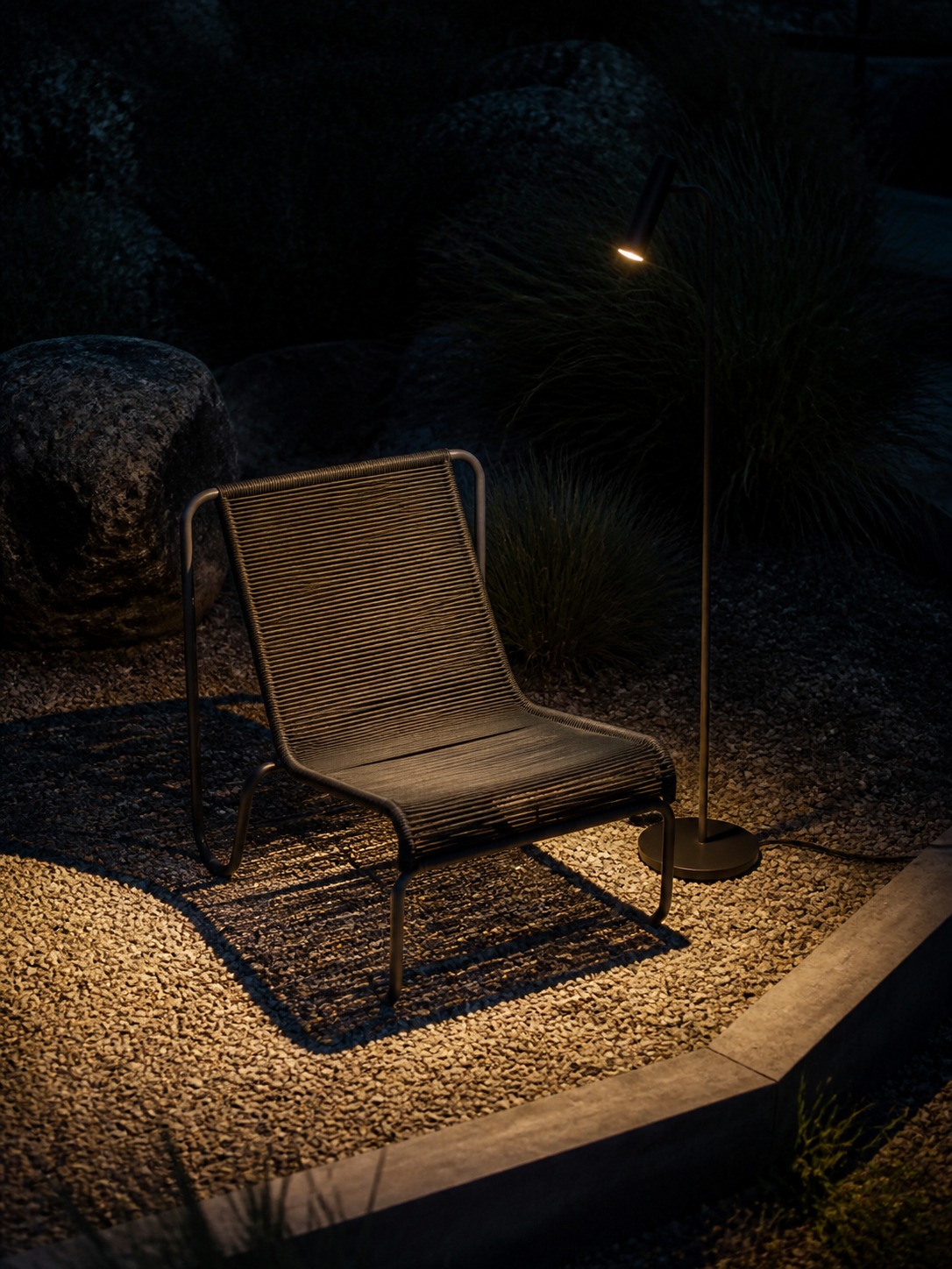 Quiet outdoor reading zone with sculptural chair A single outdoor woven chair on gravel, lit by a nearby floor lamp in a dark garden setting. | Sky Rye Design Single sculptural chair on a gravel platform with a slim floor lamp creating a quiet outdoor nook