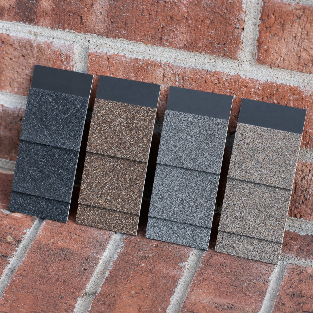 Four roof shingle color sample chips held against a red brick facade