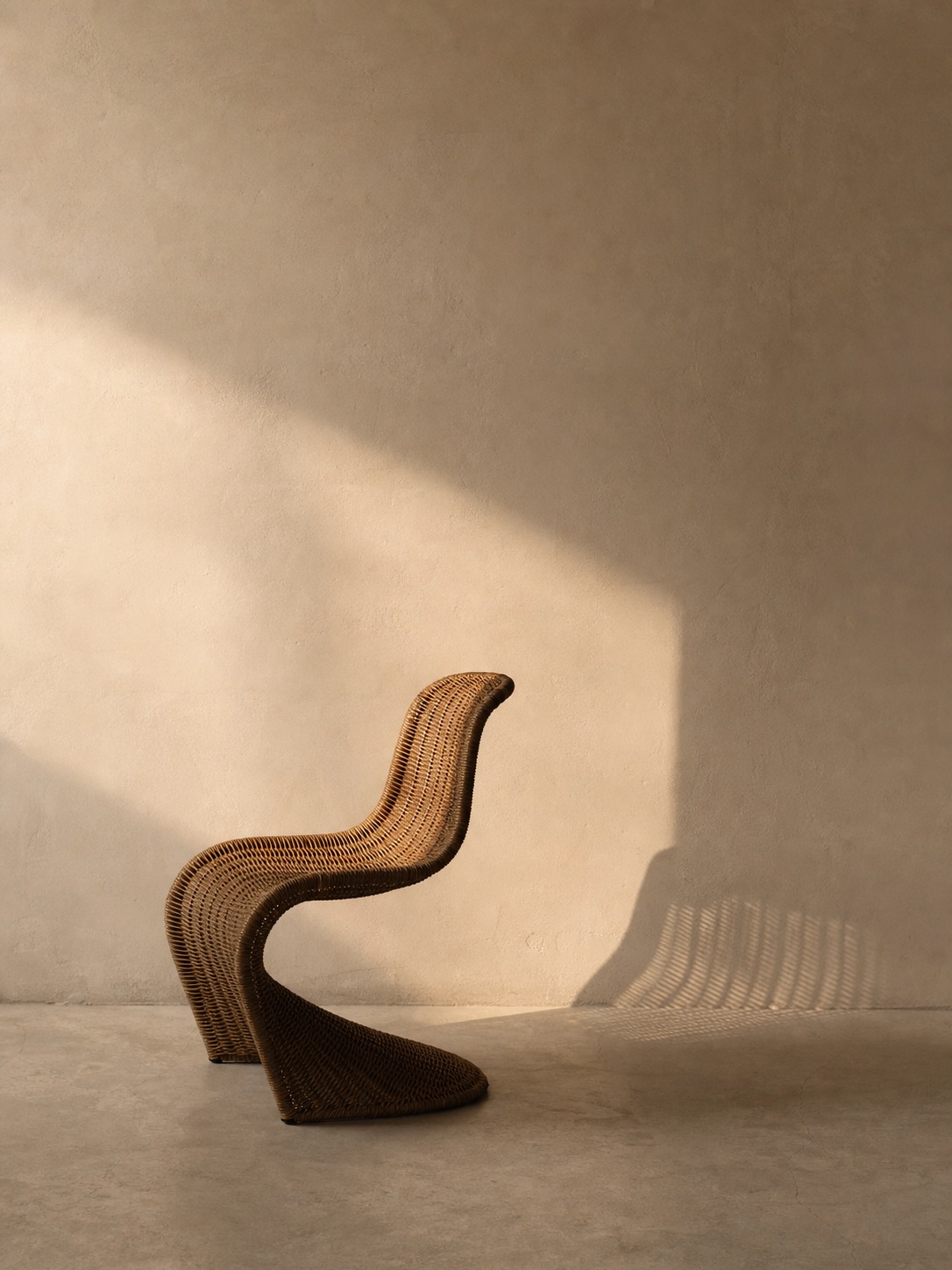 Sculptural rattan chair silhouette A sculptural rattan chair photographed alone against a plain plaster wall with soft late-afternoon shadow and generous negative space. | Sky Rye Design A sculptural rattan chair photographed alone against a plain plaster wall with soft late-afternoon shadow and generous negative space.