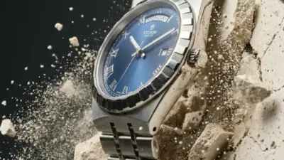 Tudor stainless steel luxury watch with blue dial and day-date display embedded in cracked stone with flying debris