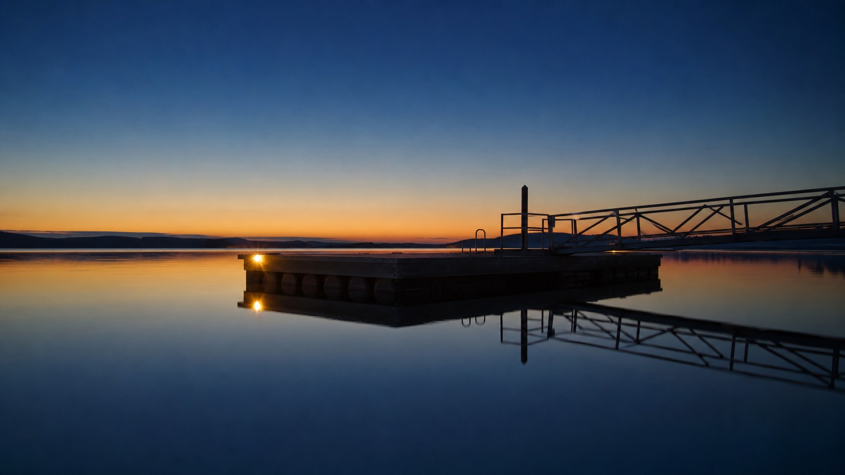 Silhouette of a low floating platform and gangway at dusk with a warm edge light reflected in still water.