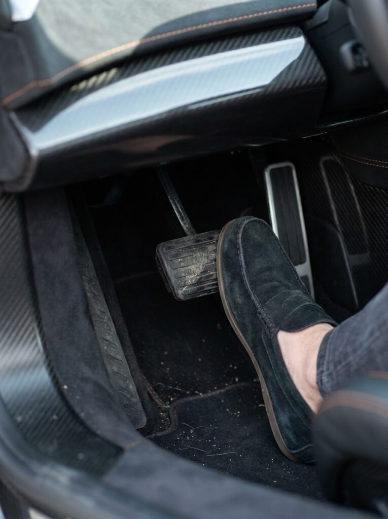 Foot_pressing_brake_pedal_202607160341 Accelerator pedal with driver foot in suede loafer inside luxury car with carbon-fiber trim | Sky Rye Design Accelerator pedal with driver foot in suede loafer inside luxury car with carbon-fiber trim