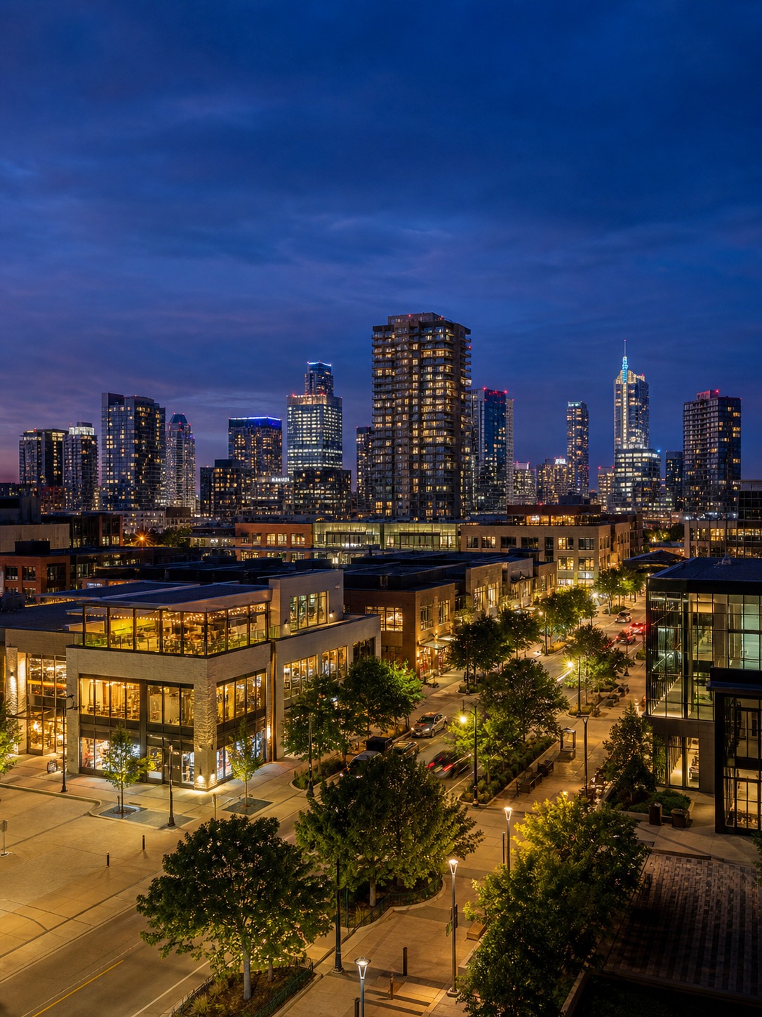 Twilight view of a walkable mixed use district with low-rise storefronts and taller towers beyond.