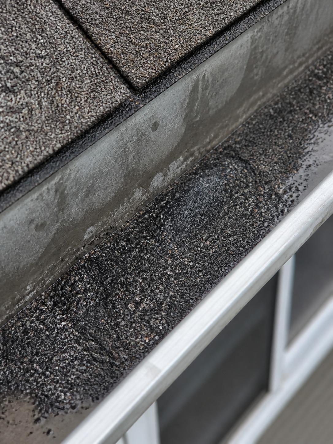 Granules in the gutter close detail Dark asphalt shingle granules collected inside a metal gutter like coarse sand. | Sky Rye Design Dark asphalt shingle granules collected inside a metal gutter like coarse sand.