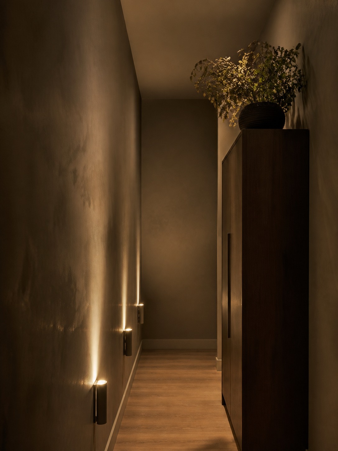 Faux plant in windowless hallway Dim, narrow hallway with warm wall lights along the left wall and a tall dark cabinet topped with a leafy potted plant on the right. | Sky Rye Design Faux architectural plant styled on top of a tall minimalist armoire in a warm windowless hallway.