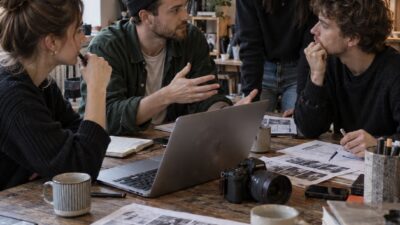 Four people collaborate around a laptop on a cluttered wooden table in a bright studio, reviewing photographs.