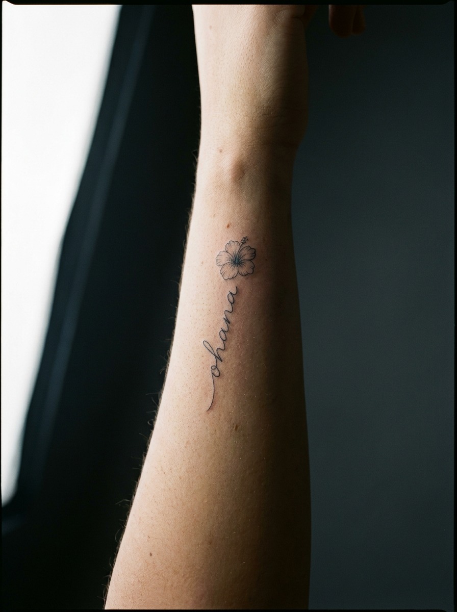 Raised forearm with ohana script and a small hibiscus accent tattoo.