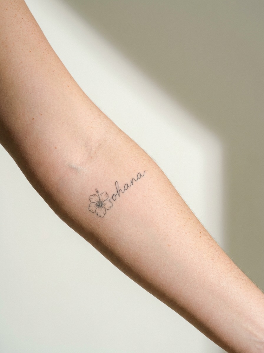 Forearm tattoo with cursive ohana script and a small hibiscus bloom beside it.