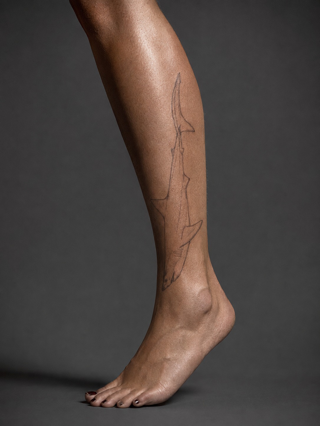 Model showing a fine-line mako shark tattoo wrapping the calf with realistic healed ink and editorial lighting.