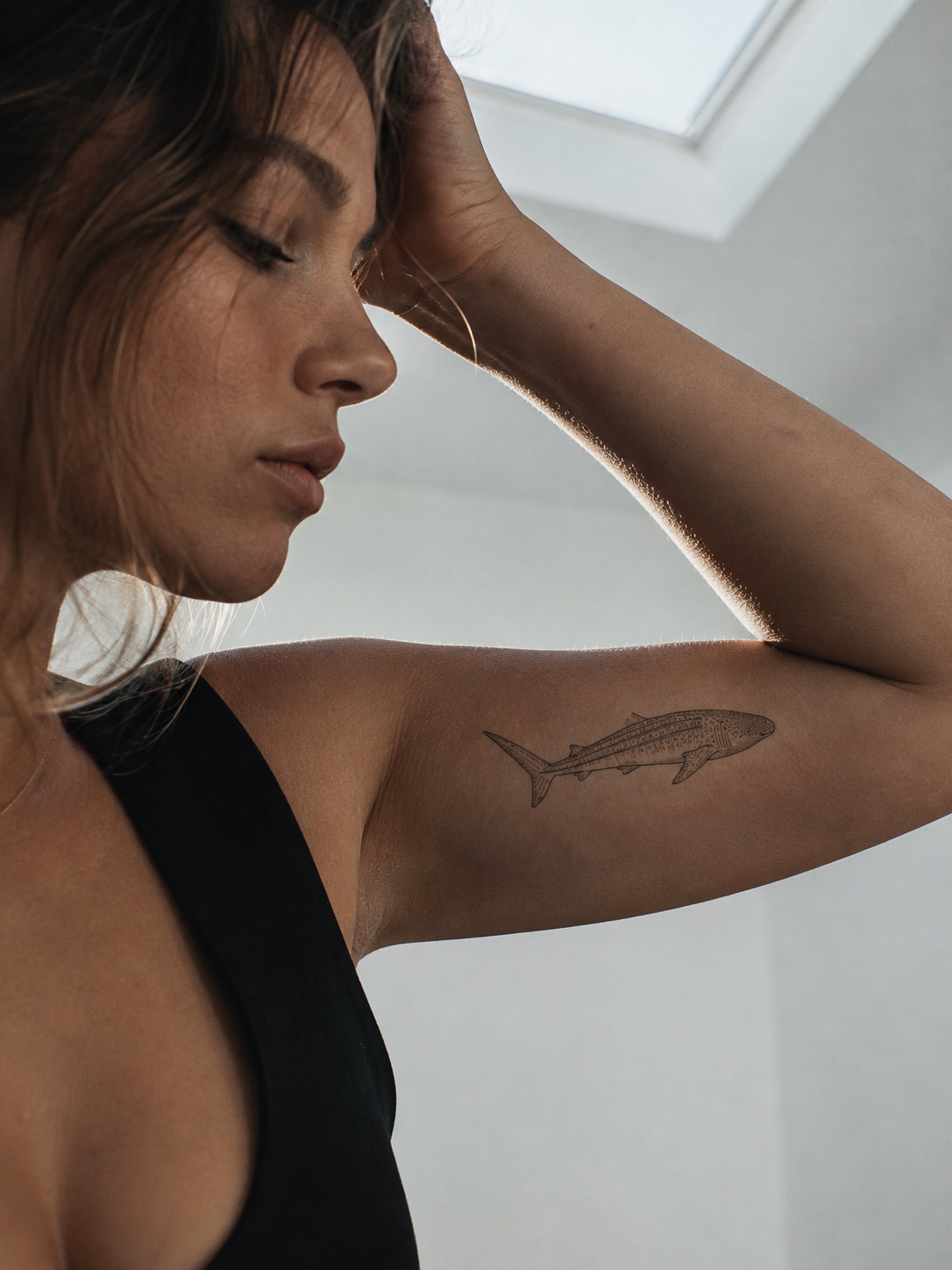 Model showing a simplified fine-line whale shark outline tattoo on the inner bicep with realistic healed ink and editorial lighting.