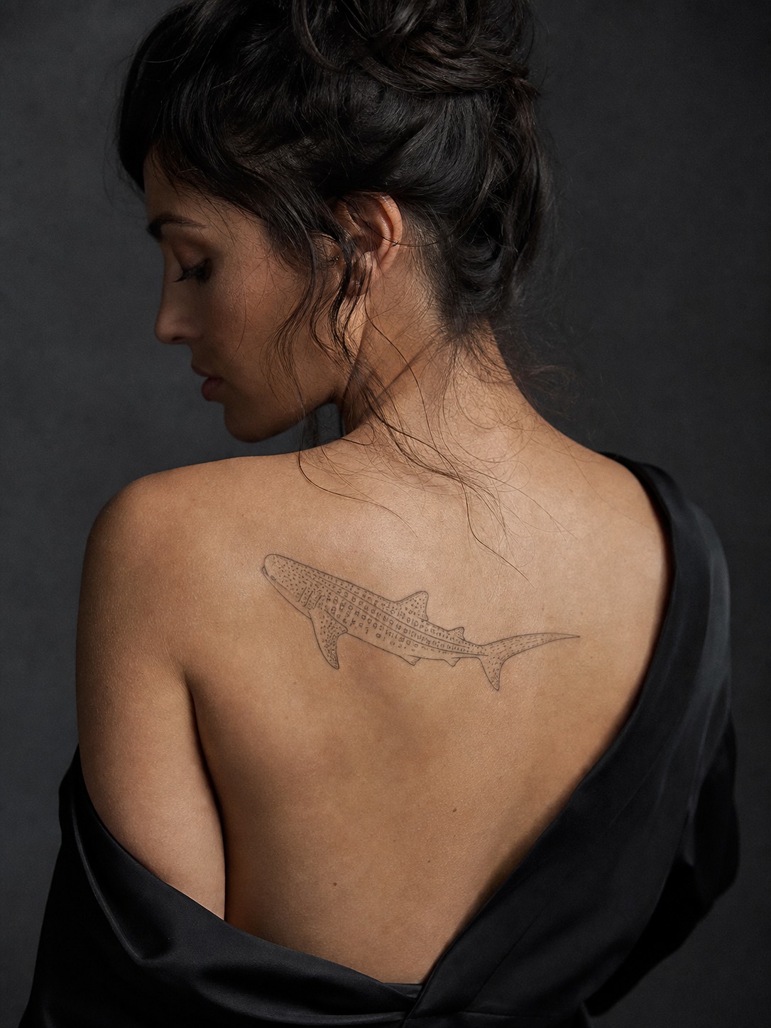 Model showing a simplified outline whale shark tattoo across the shoulder blade with realistic healed ink and editorial lighting.