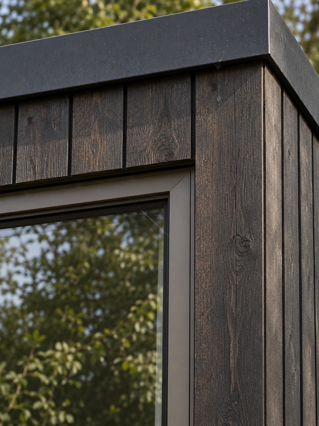Close detail where dark stained timber cladding meets a matte metal window frame and the roof edge