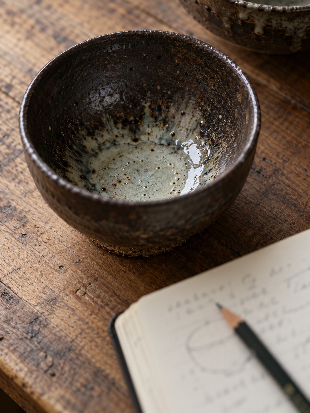 Describe it then explain it Close-up of pale ash glaze pooling inside a dark stoneware tea bowl, with a notebook and pencil blurred beside it | Sky Rye Design Close-up of pale ash glaze pooling inside a dark stoneware tea bowl, with a notebook and pencil blurred beside it