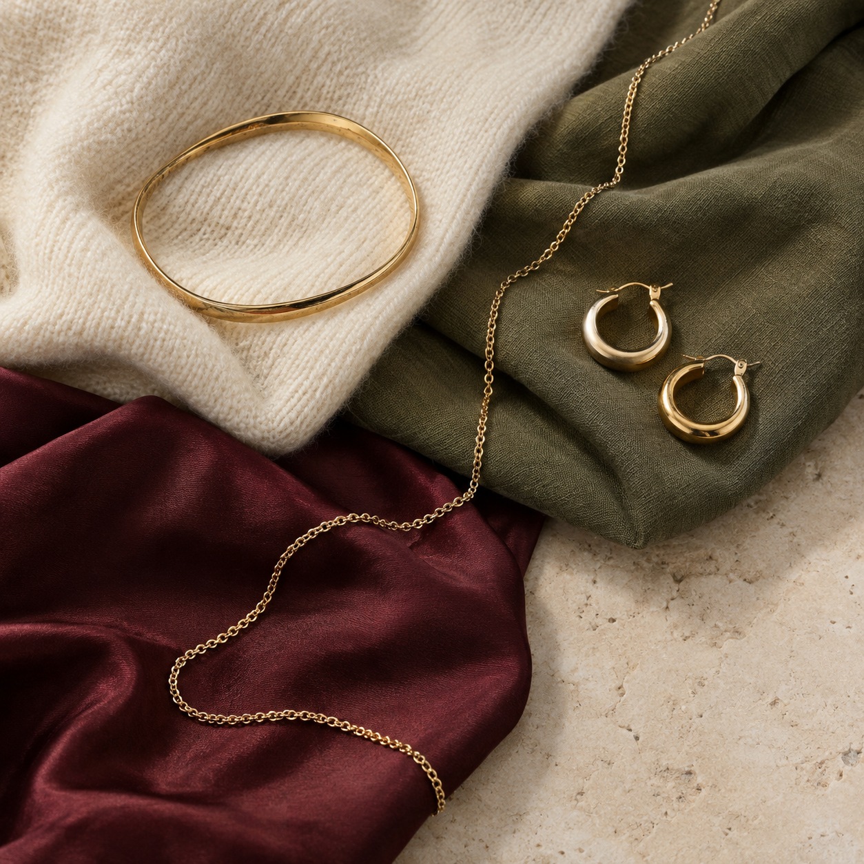 Gold jewelry with warm wardrobe colors Gold-plated bracelet, hoops and chain arranged across cream wool, muted olive cotton and burgundy silk. | Sky Rye Design Gold-plated bracelet, hoops and chain arranged across cream wool, muted olive cotton and burgundy silk.