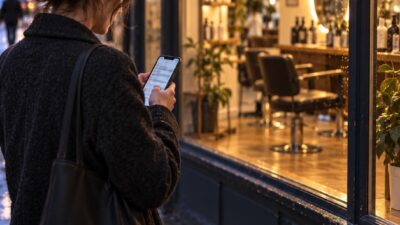 A client uses a phone outside a warmly lit salon at dusk.